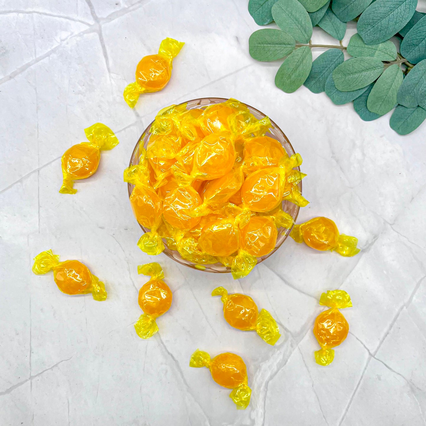 Yellow candies in a glass bowl on a white surface with green leaves.
