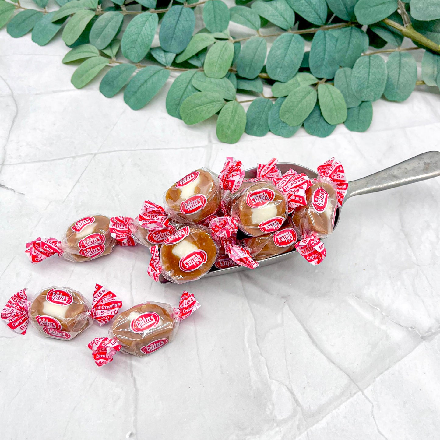 Spoonful of wrapped caramel cream candies with red and white wrappers on a light background with green leaves.