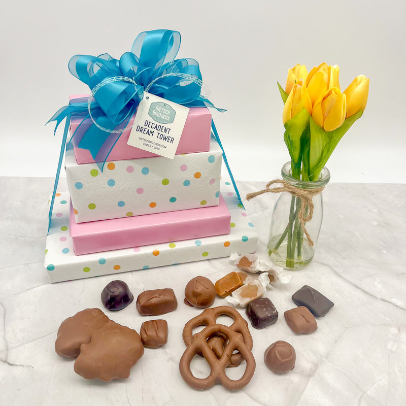 Four layered tower of assorted treats wrapped in multi colored polka dots on white paper and solid light pink wrapping paper and tied with a blue bow.