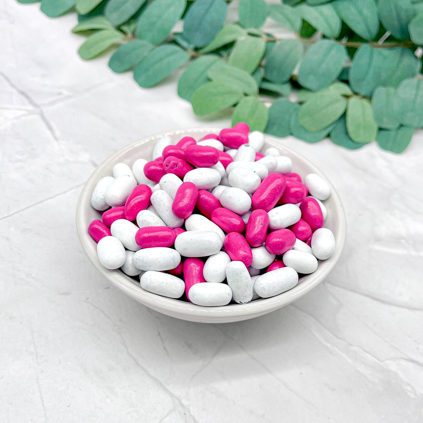 White bowl filled with pink and white candies on a light gray surface with green leaves in the background