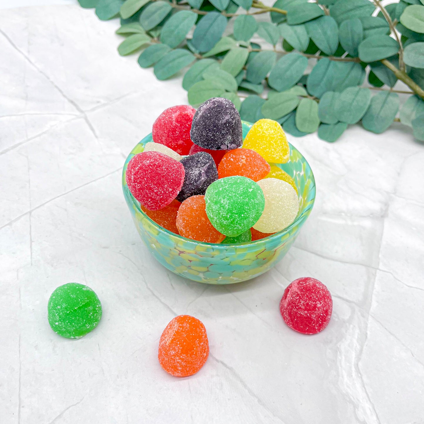 Colorful gummy candies in a small bowl on a white surface with green leaves in the background