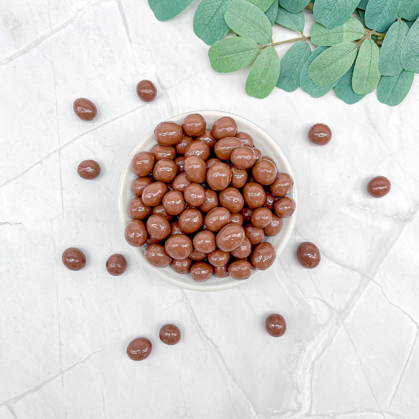 Chocolate-covered espresso beans in a white bowl on a light background with green leaves.