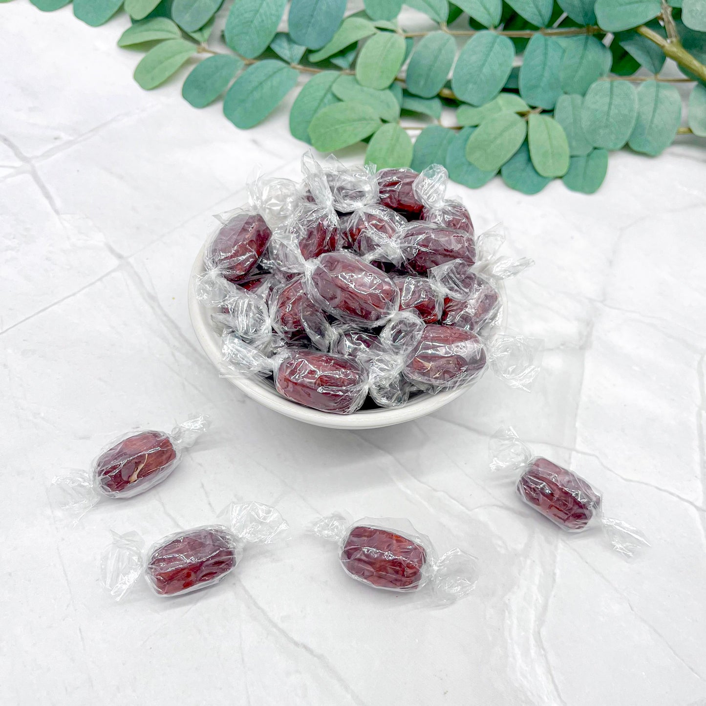 Root beer barrels in clear wrappers on a white surface with green leaves in the background