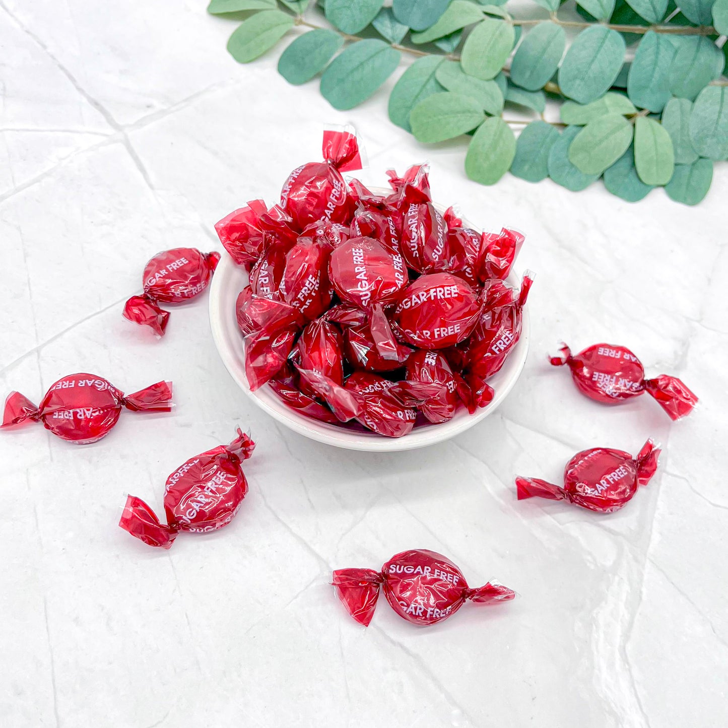 Red sugar-free candies in a white bowl on a light surface with green leaves.