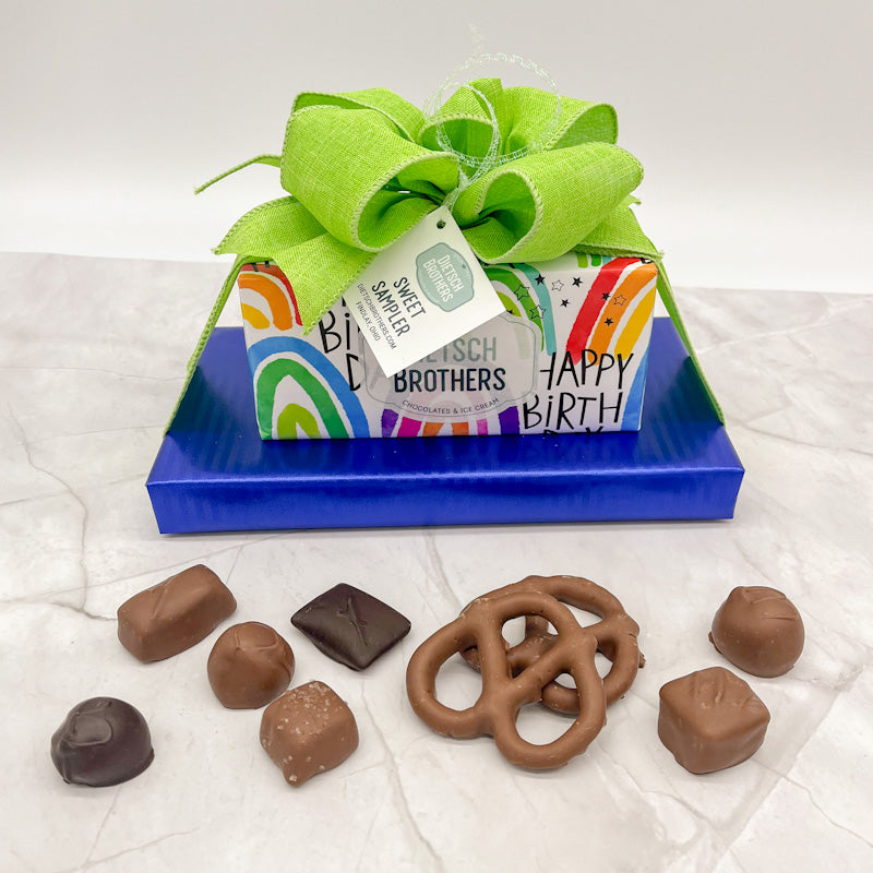Two layered tower of assorted treats wrapped in multi colored Happy Birthday paper and solid dark blue paper and topped with a bright green bow.