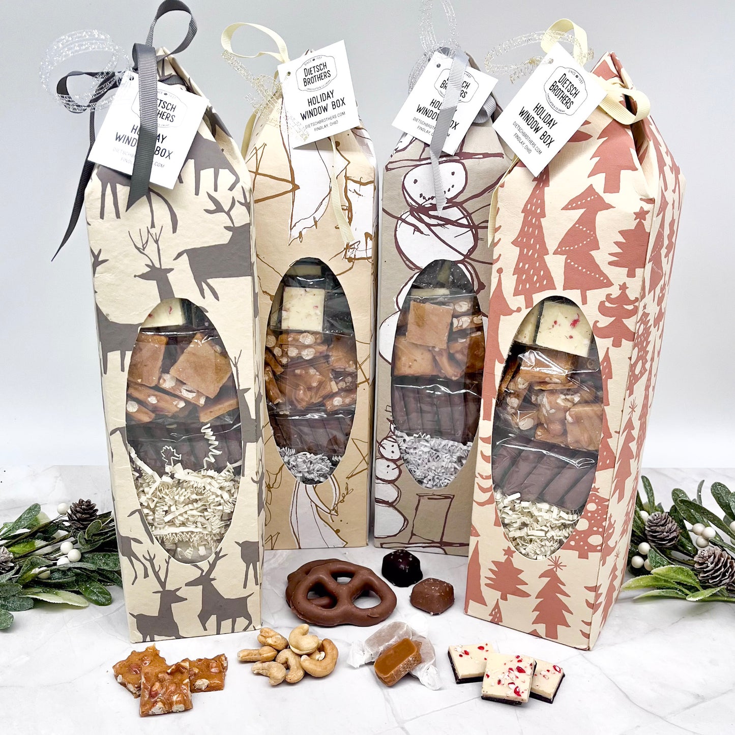 Four gift boxes with festive designs and treats on a white surface with greenery.