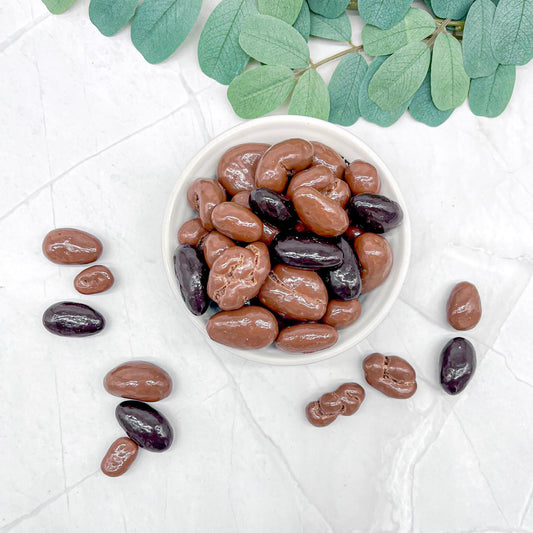 Bowl of milk and dark chocolate-covered nuts on a white surface with green leaves.