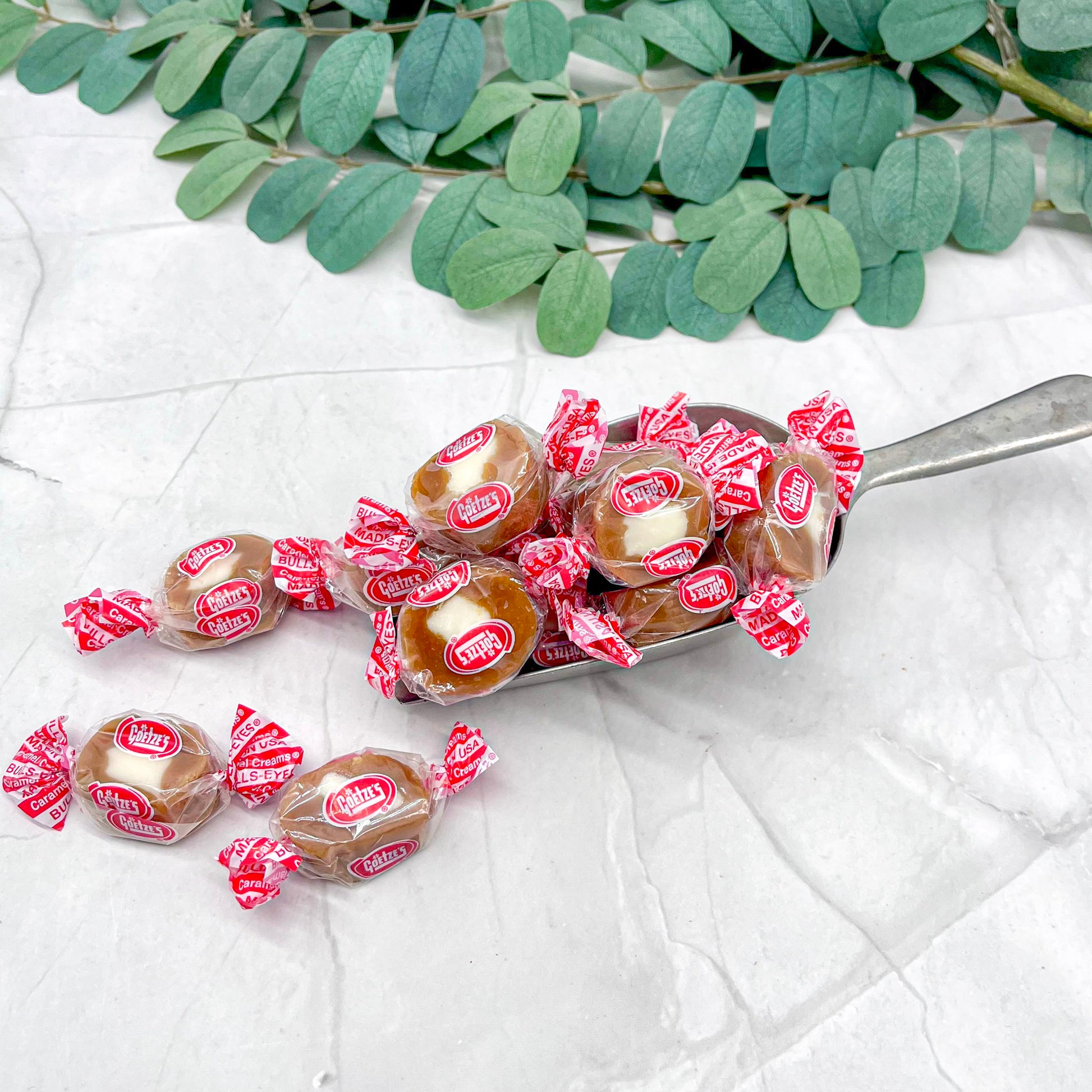 Spoonful of wrapped caramel cream candies with red and white wrappers on a light background with green leaves.