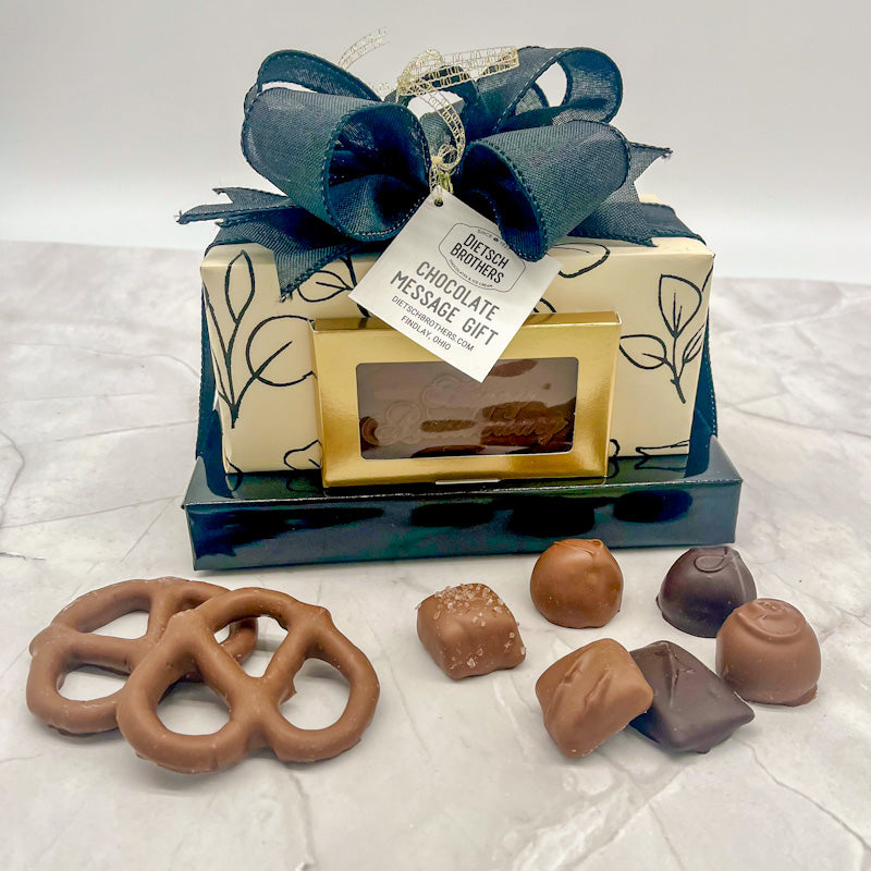 Two layered tower of assorted treats wrapped in solid navy blue paper and black florals on cream paper, with a milk chocolate bar featuring "Happy Anniversary" on the front of the package, all topped with a navy blue bow