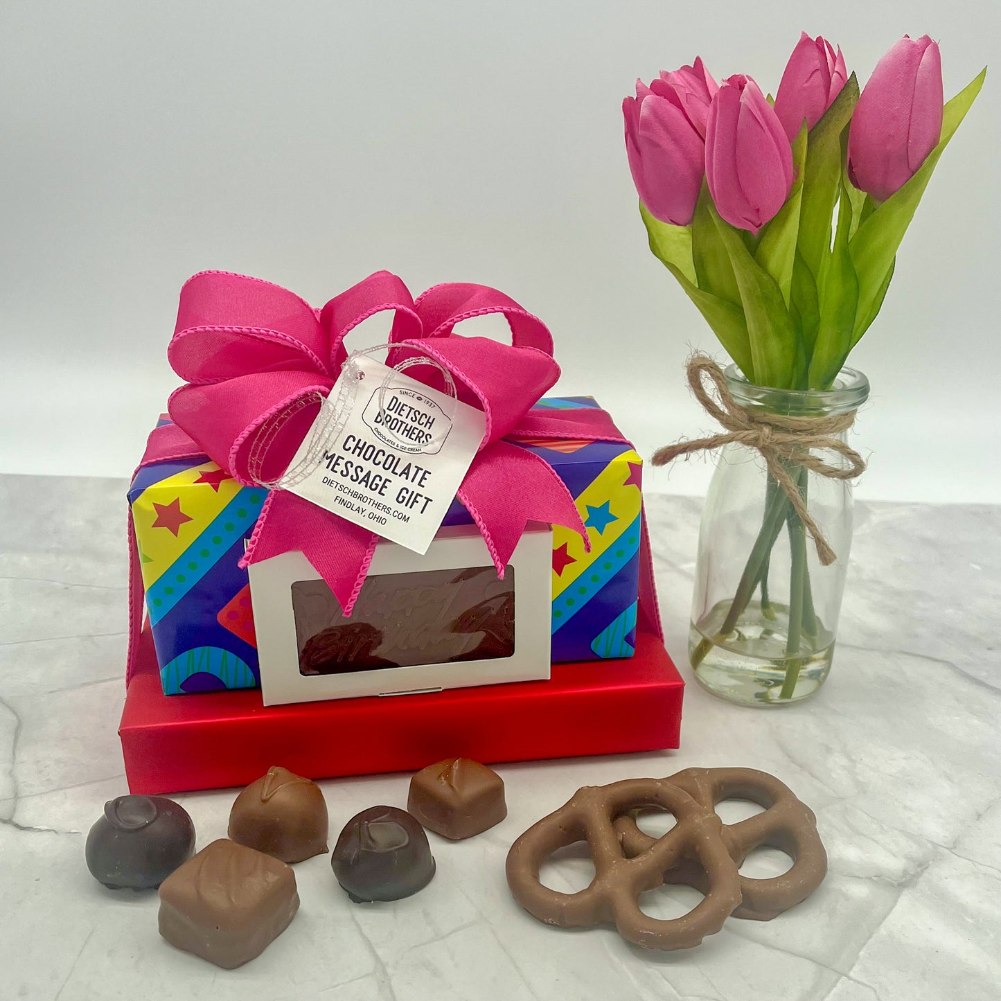 Two layered tower of assorted treats wrapped in solid red paper and "happy birthday" paper, with a milk chocolate bar featuring "Happy Birthday" on the front of the package, all topped with a pink bow
