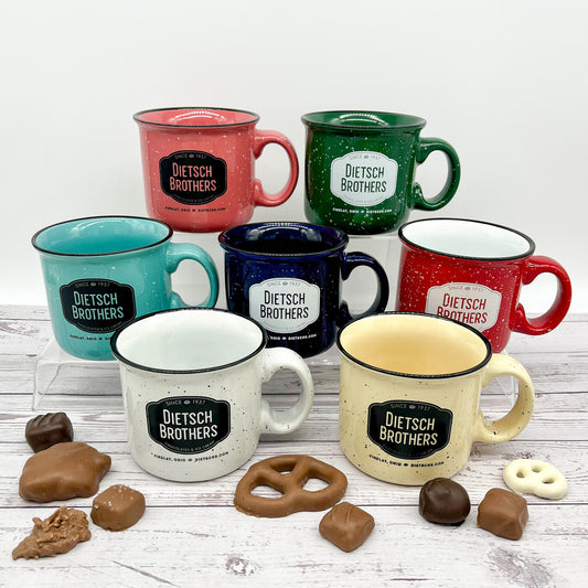 Collection of Dietsch Brothers mugs in various colors on a wooden surface with candies and pretzels sprinkled around it.