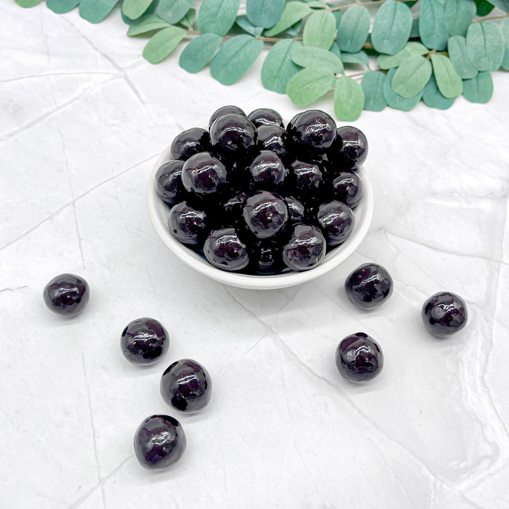 Dark chocolate covered malt balls in a white bowl on a light surface with green leaves.