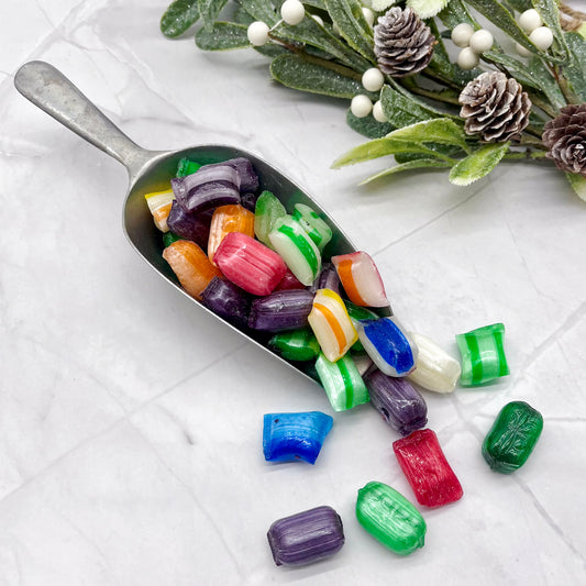 Metal scoop filled with colorful candies on a white surface with decorative greenery.