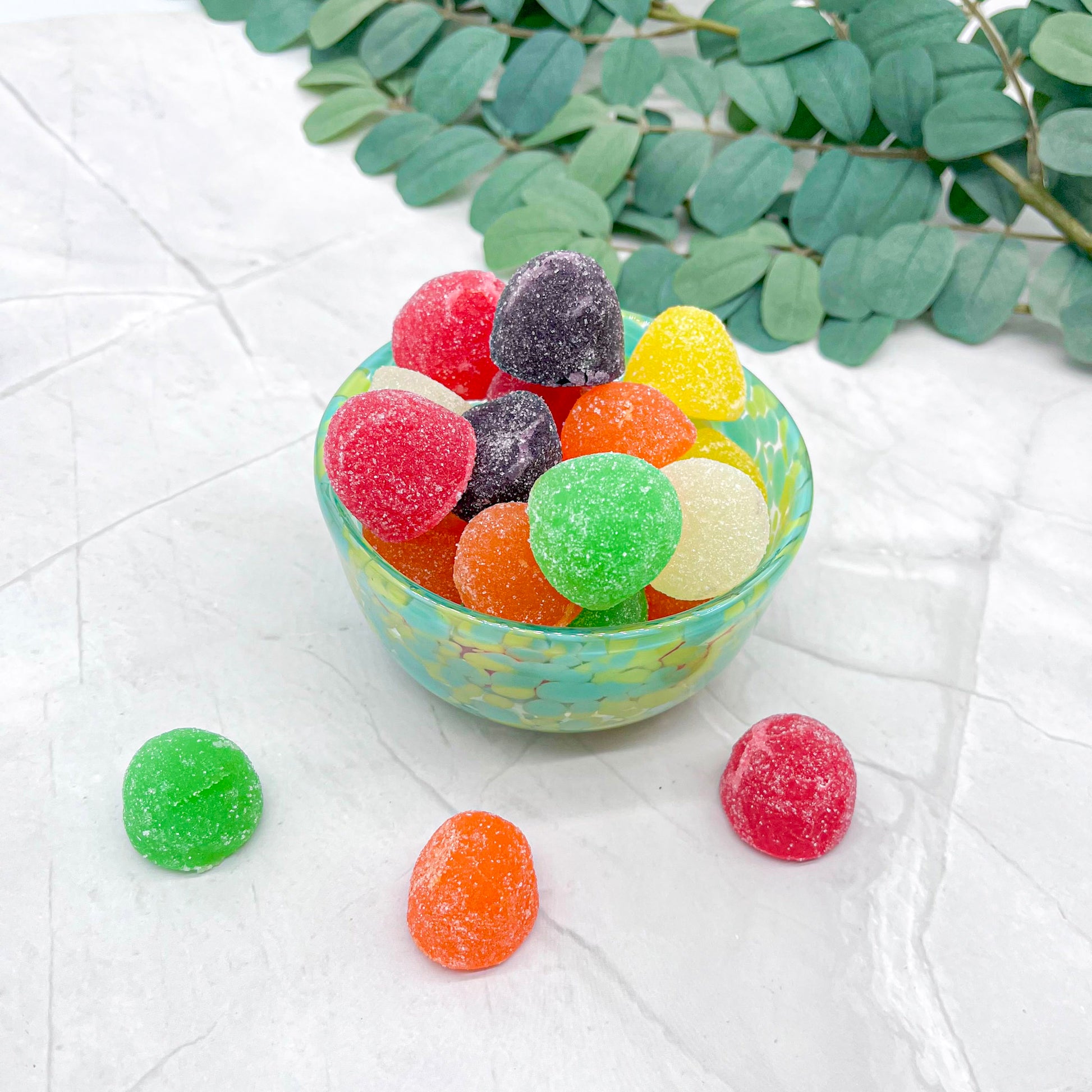 Colorful gummy candies in a small bowl on a white surface with green leaves in the background