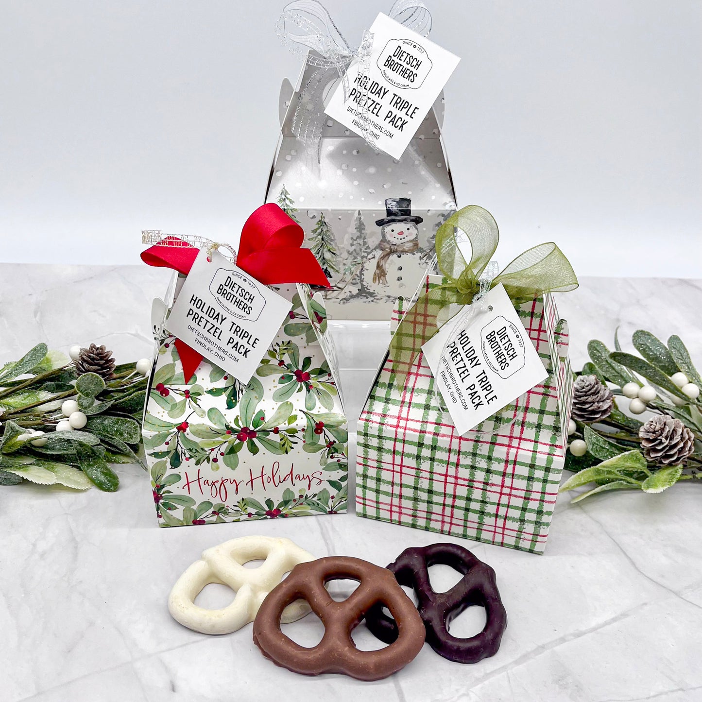 Holiday-themed pretzel packs with decorative ribbons and tags on a white surface with greenery.