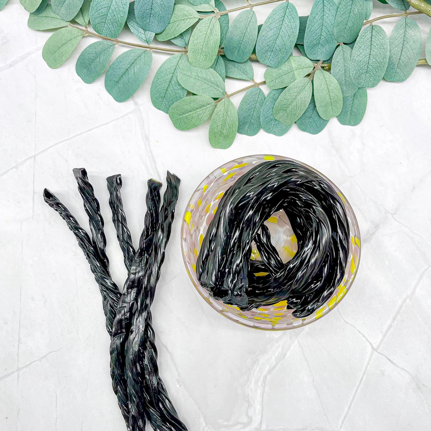 Black licorice sticks and a glass container with more licorice on a white surface with green leaves.