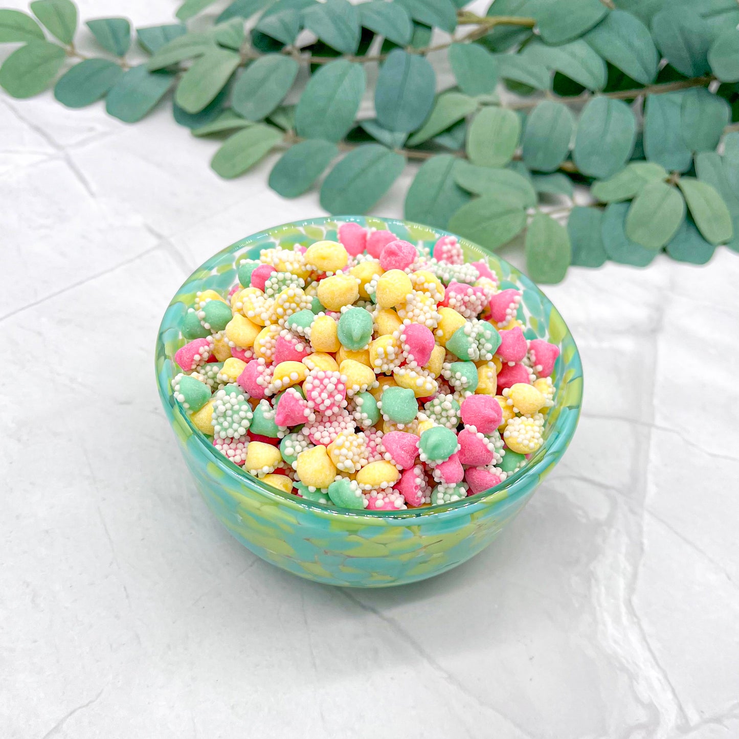 Small bowl filled with colorful pastel mini mints with white sprinkles on a light surface with green leaves in the background
