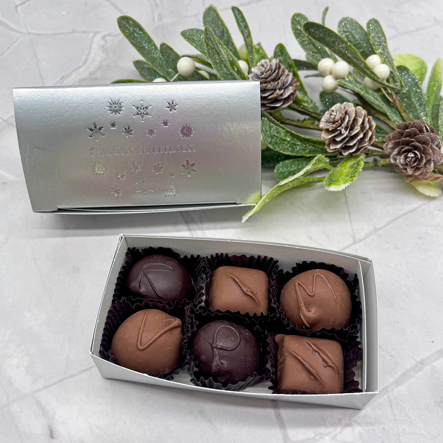 Box of chocolates with a decorative silver cover on a textured surface with greenery.
