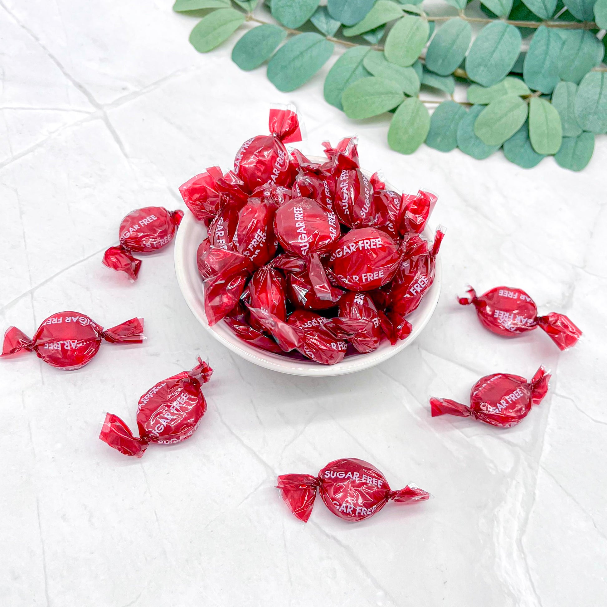 Red sugar-free candies in a white bowl on a light surface with green leaves.