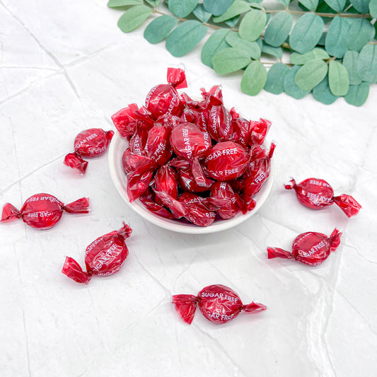 Red sugar-free candies in a white bowl on a light surface with green leaves.