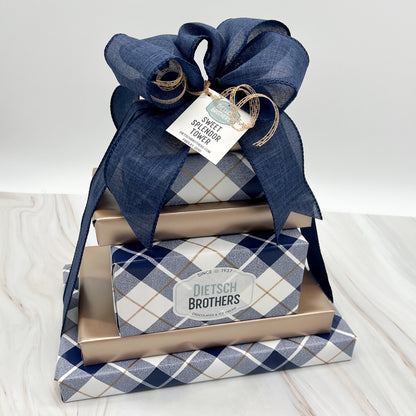 Six layered tower of assorted treats wrapped in navy and white plaid paper and solid light copper wrapping paper and tied with a navy blue bow.